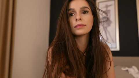 Video of a young woman with long, straight brown hair, wearing a sleeveless brown top, standing indoors. Background features a black-framed monochromatic portrait and beige curtains.