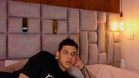 Media: Video of a young Hispanic man with short dark hair, lying on a bed with a gray padded headboard, wearing a black t-shirt, and looking contemplative.