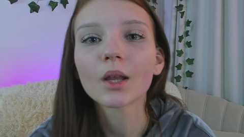 Media: Video of a young Caucasian woman with fair skin, long brown hair, and light makeup, wearing a gray shirt, in a cozy indoor setting with soft purple lighting and green star garland.