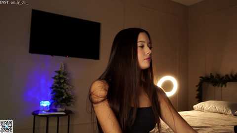 Media: Video of a young, slim, Latina woman with long, straight black hair, wearing a black strapless top, standing in a dimly lit bedroom with a glowing round mirror, Christmas tree, and wall-mounted TV.