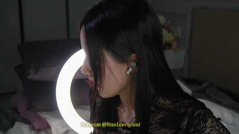 Media: Video of a woman with long, straight black hair wearing a black lace top, leaning on a bed with a large circular ring light. Background includes a white wall, a vase with flowers, and a mirror.