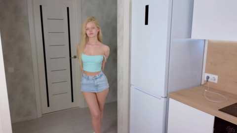 Media: A video of a slender, blonde woman in a strapless, light blue top and denim shorts standing in a modern, minimalist kitchen with white cabinets and light-colored countertops.