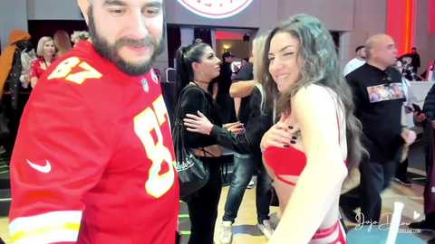 Media: Video of a smiling, curvy brunette woman in a red sports bra and black shorts, standing with a bearded man in a red jersey at a crowded indoor event.