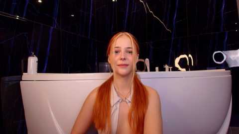 Video of a young, fair-skinned woman with long, straight red hair, sitting in a modern, white bathtub. The background features dark, abstract shapes and a minimalist design.