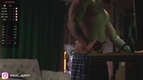 Video of a shirtless man with a muscular build, wearing plaid pajama bottoms, standing in a dimly lit room with a green screen in the background.