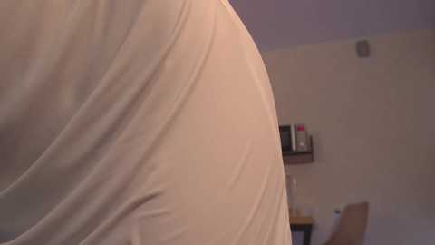 Media: A close-up video of a person's upper torso, wearing a white shirt, in a dimly lit room with beige walls, a wooden table, and a chair.