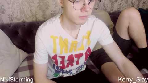 Media: Video of a pale-skinned, androgynous person with glasses, wearing a white \"Treat\" graphic tee and black shorts, seated on a purple velvet sofa with patterned wallpaper in the background.