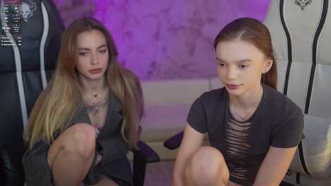 Video of two young women with long hair, sitting on the floor. One has a tattoo, wearing a gray top; the other has a striped shirt. Purple lighting, indoor setting.