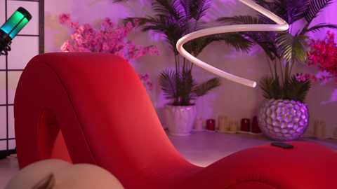 Media: Video of a modern, red chaise lounge with a white, spiral-shaped lamp in a room with pink flower wallpaper, greenery, and candles on a table.