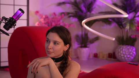 Media: Video of a nude woman with long brown hair, sitting on a red couch, wearing a black choker, and holding a microphone, in a modern, dimly lit room with a white backdrop and purple lighting.