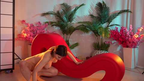 Media: Video of a nude woman with dark hair and medium breasts, kneeling on a red, curvy chaise lounge in a room with green plants, pink flowers, and a black room divider.