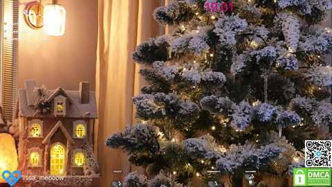 Media: A cozy video of a warmly lit living room with a snow-covered Christmas tree adorned with twinkling lights. A wooden house decoration with glowing windows stands beside it, against a beige curtain backdrop.