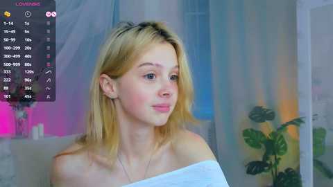 Media: A video of a young, fair-skinned blonde woman with shoulder-length hair, wearing a white off-shoulder top, sitting indoors with a soft, pastel-colored backdrop.