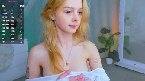 Media: Video of a young, fair-skinned woman with long, wavy blonde hair, wearing a white shirt, partially covering her breasts, in a minimalist room with green plants.