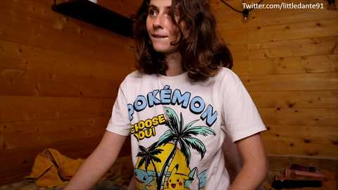 Media: Video of a young woman with shoulder-length curly brown hair, wearing a \"Pok\u00e9mon\" T-shirt, in a wooden-paneled room.