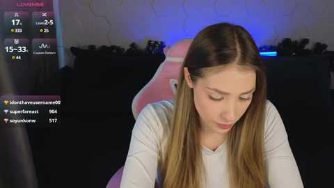 Video of a young woman with long, straight, light brown hair, wearing a white long-sleeved shirt, sitting in a pink gaming chair, in a dimly lit room with a blue light on the wall.