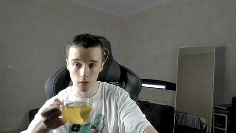 Video of a young man with short dark hair, wearing a white T-shirt, sitting in a black gaming chair. He holds a glass of orange juice. Background features a plain wall, a desk with a monitor, and a mirror.