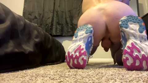 Media: Video of a nude man with fair skin, muscular build, and visible circumcised penis, kneeling on a beige carpet, wearing vibrant sneakers. Background shows a dark blanket and gray curtain.