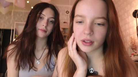 Media: Video of two young women with long hair, one with a nose ring, both wearing casual tops, in a cozy, softly lit room with a warm, inviting atmosphere.