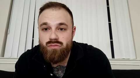 Media: Video of a bearded man with short brown hair, wearing a black shirt, indoors with white vertical blinds in the background.