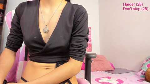 Media: Video of a woman with light brown skin, wearing a black cropped top and black panties, seated in a pink gaming chair. Background features a bed with pink sheets and a laptop.