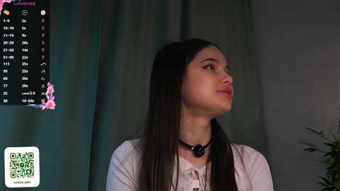 A video of a young woman with long dark hair and fair skin, wearing a white top and black necklace, standing against a dark curtain.