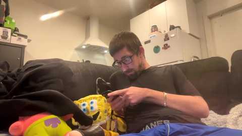 Media: Video of a man with glasses and a beard, wearing a black shirt and blue pants, playing a video game on a couch, surrounded by stuffed toys and kitchen appliances in a cozy, cluttered living room.