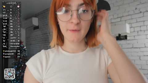 Video of a young woman with fair skin and red hair, wearing glasses and a beige top, adjusting her hair in a modern, dimly lit room with a white brick wall.