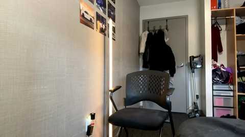 Media: Video of a sparsely furnished, minimalist bedroom with light grey walls, a black folding chair, a tall lighted mirror, a closed grey door, and a wooden closet with hanging clothes and laundry.