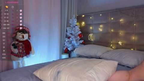 A cozy bedroom photo featuring a plush dog toy wearing a festive scarf, a Christmas tree with lights, and a bed with white pillows and a gray headboard.