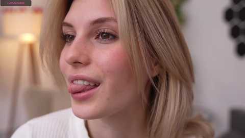 Media: Video of a fair-skinned, blonde woman with shoulder-length hair, slightly parted lips, and a neutral expression. She wears a white top. Background is blurred, showing a lamp and wall decor.