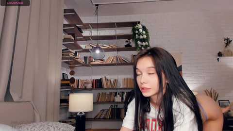 Media: Video of an Asian woman with long black hair, wearing a white T-shirt, sitting on a bed in a cozy, bookshelf-lined room with a wooden ceiling and hanging pendant light.