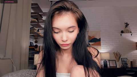 Media: A video of a young woman with long, straight black hair, fair skin, and closed eyes, sitting in a cozy, modern bedroom with a white brick wall and minimalist decor.