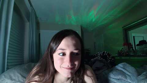 Media: Video of a young woman with long brown hair, smiling, in a dimly lit bedroom with a green laser light display on the ceiling.