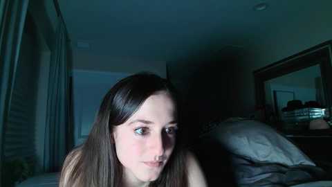 Media: Video of a young Caucasian woman with long brown hair, pale skin, and freckles, wearing a sleeveless top, sitting on a bed with rumpled white sheets in a dimly lit room.