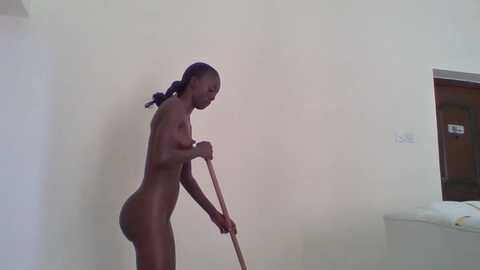 Media: Video of a nude, dark-skinned woman with braided hair, scrubbing a white wall with a mop. She stands in a simple, sparsely furnished room with a bed and a door.