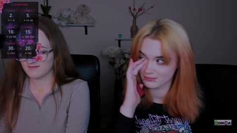 Media: Video of two women in a dimly lit room: one with long, straight brown hair, wearing glasses and a light grey sweater, the other with shoulder-length, vibrant orange hair, a black shirt, and a phone to her ear.