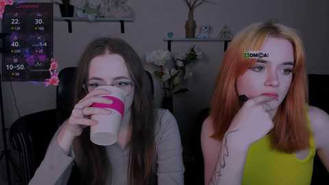 Media: Video of two young women, one with long brown hair, the other with orange hair, sitting side by side, drinking from cups, in a dimly lit room with a digital clock and floral decor.