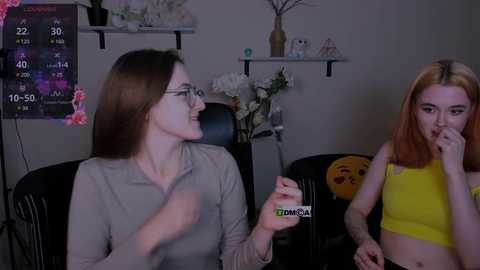 Media: Video of two women, one with glasses, gray top, and light skin, and the other with red hair, yellow crop top, and fair skin, sitting on black chairs, in a dimly lit room with floral decor and a clock.