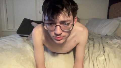 Video of a young, fair-skinned, shirtless man with short, dark hair and glasses, lying on a bed with a beige blanket, in a dimly lit, modest bedroom setting.