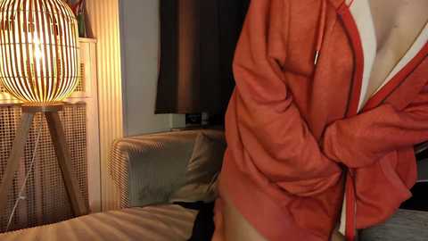 Media: A video of a person in an orange jacket, holding the jacket open, standing in a dimly lit bedroom with a modern lamp and a dark TV screen in the background.