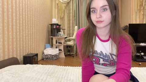 Media: Video of a young Caucasian woman with light skin and long brown hair, wearing a pink and white long-sleeve shirt, sitting on a bed in a cluttered, beige wallpapered room.