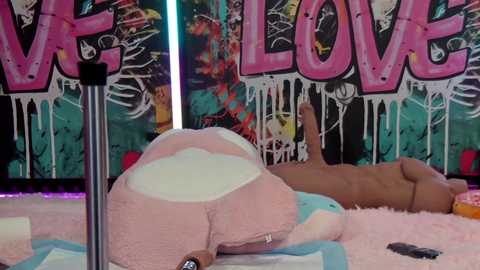 Video of a plush bunny and a nude male figure lying on a pink, fluffy rug in a graffiti-covered room with \"WELCOME\" in bold pink letters.