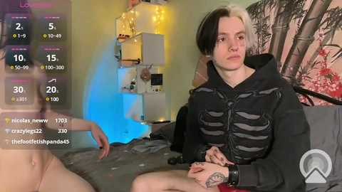 Media: A video of a pale-skinned, androgynous individual with short, dyed hair, wearing a black hoodie, seated on a bed with a skeletal motif. The background features a colorful, anime-style poster and a yellow lamp.