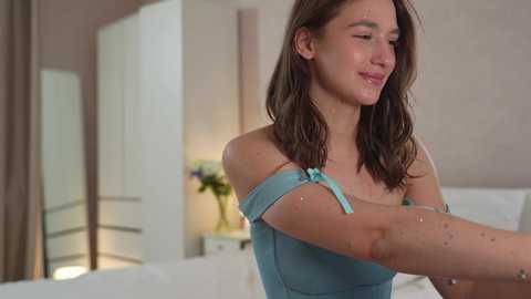 Media: Video of a young Caucasian woman with wavy brown hair, wearing a light blue off-shoulder top, standing in a modern bedroom with white furniture and beige walls.