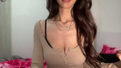 Media: Video of a woman with long, wavy brown hair, wearing a beige ribbed top with a black bra, silver necklace, and pink bedspread in a bedroom.