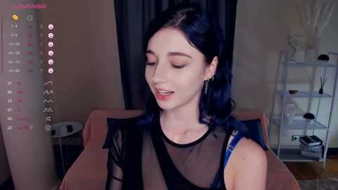 Video of a young woman with fair skin and long, dark hair, wearing a sheer black top, sitting in a room with beige walls, a gray curtain, and a white shelf with decor items.