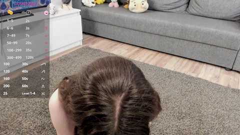 Media: Video of a person with short, voluminous, dark brown hair, sitting on a grey sofa with stuffed toys and a TV in the background.