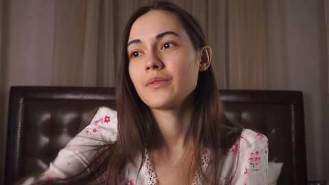 Media: Video of a young woman with long brown hair, wearing a floral-patterned blouse, sitting on a brown leather couch. She has a serene expression and is looking to the side.