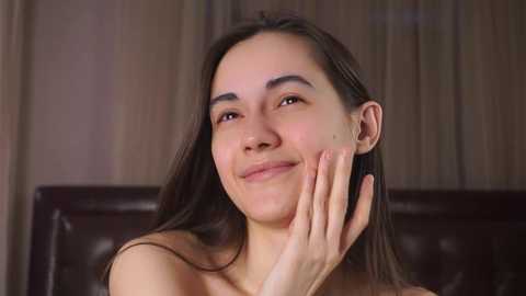 Media: Video of a smiling, light-skinned Asian woman with long dark hair, touching her face with her left hand. She's topless, wearing a dark leather headboard in the background.